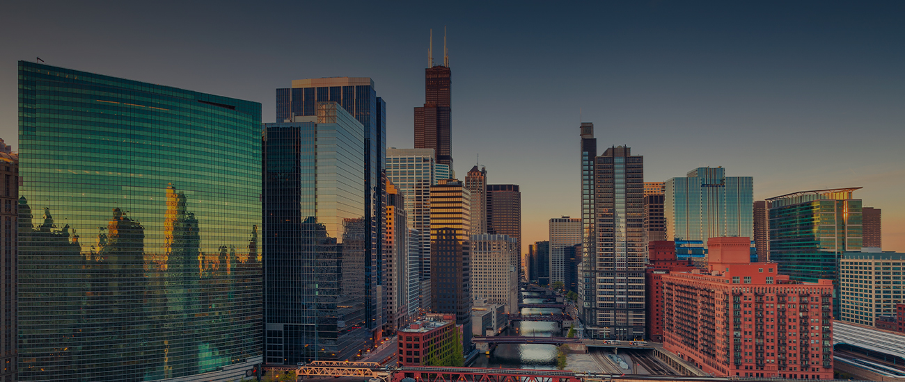 A panoramic view of a city skyline during sunset, showcasing modern skyscrapers with the prominent tower having a reddish-brown hue. The buildings reflect the golden light of the setting sun against a clear blue sky.
